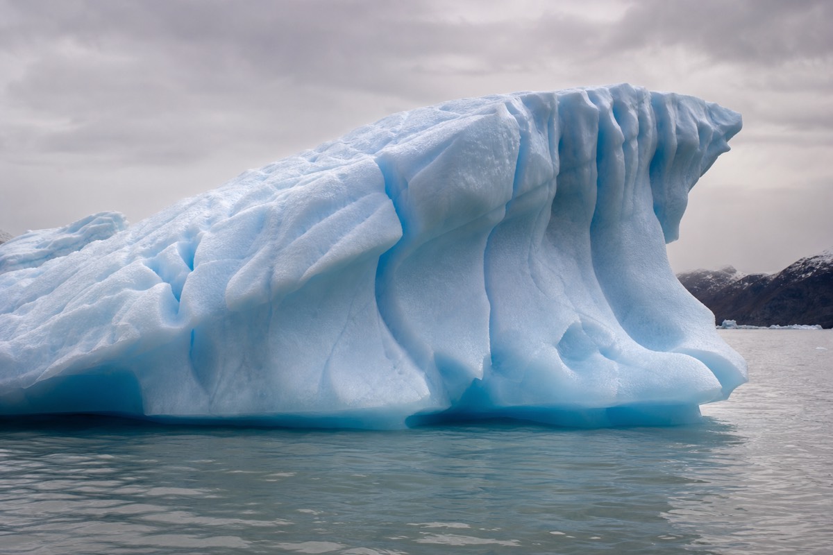 A weathered blue iceberg floating close to the surface of a grey glacial lake